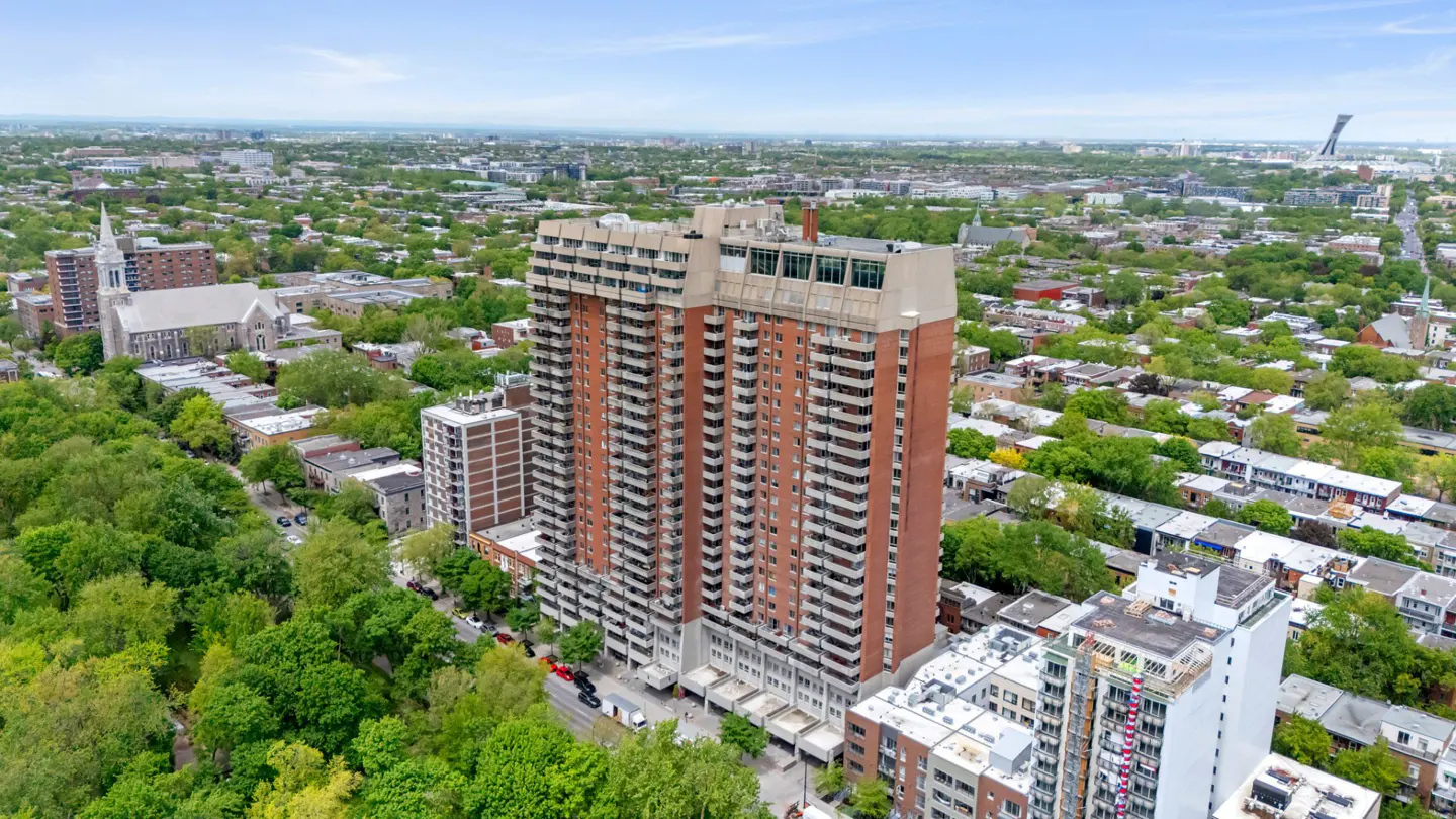 Aerial view of a tall red-brick apartment building surrounded by lush green trees and a dense urban neighborhood.