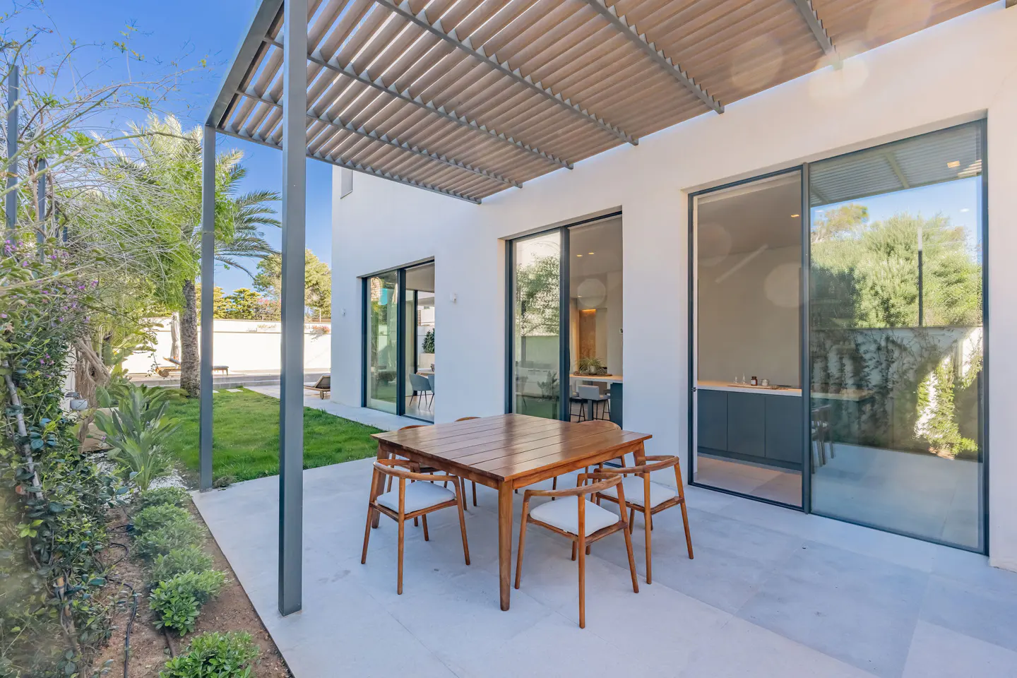 Outdoor patio with a wooden table and chairs under a pergola, next to a white building with sliding glass doors. Lush green lawn and garden visible.