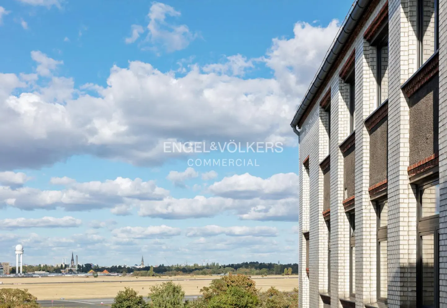 Exterior view of a white brick building with windows, against a blue sky with clouds and a distant cityscape.