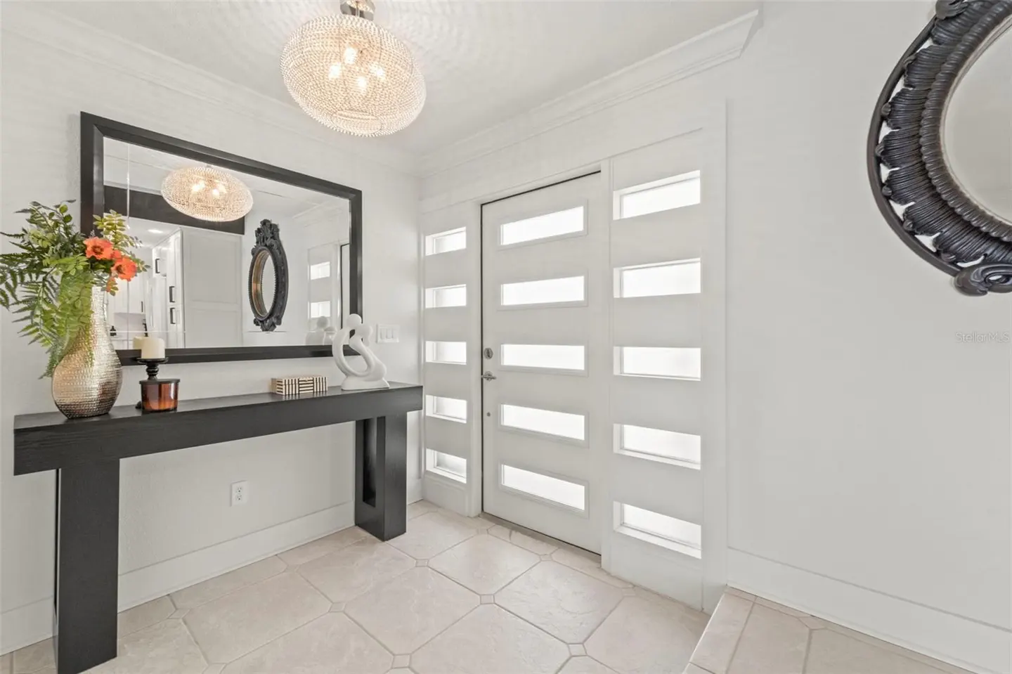 Bright foyer with white walls, a black console table with flowers, and a modern white door with glass panels. A chandelier hangs from the ceiling.