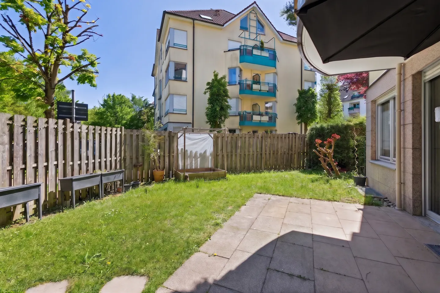 A fenced backyard with grass, a patio, and a view of a yellow apartment building with blue balconies.