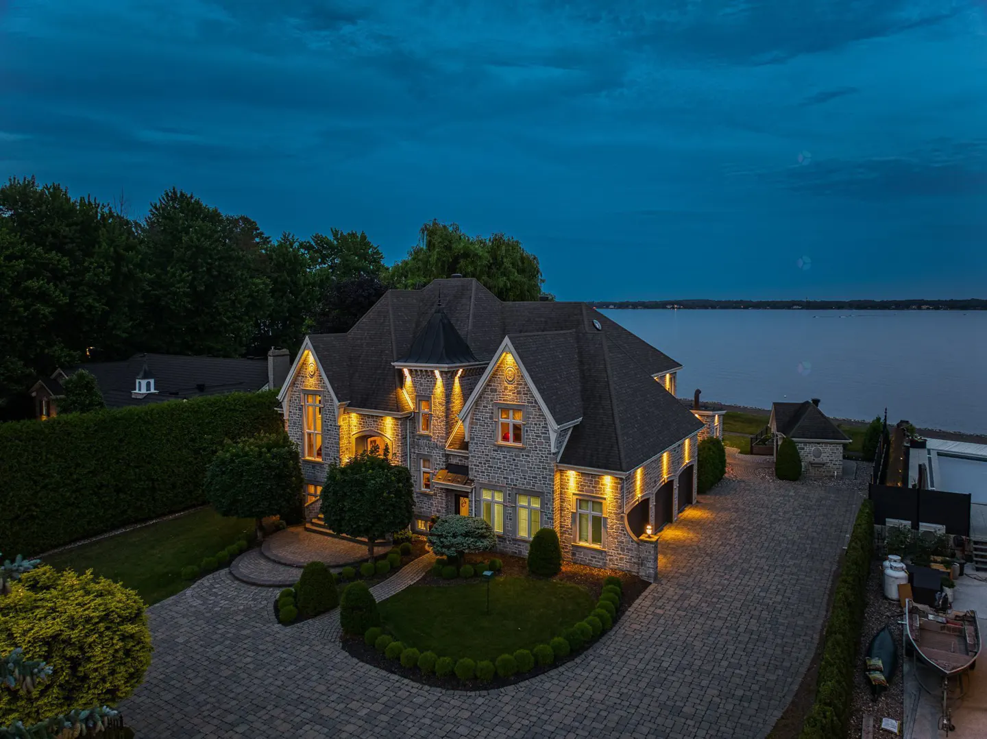 Aerial view of a large, illuminated stone house with a dark roof, green lawn, and lake background at dusk.
