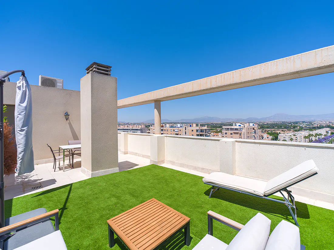 Rooftop terrace with artificial grass, a lounge chair, outdoor dining set, and panoramic city views under a clear blue sky.