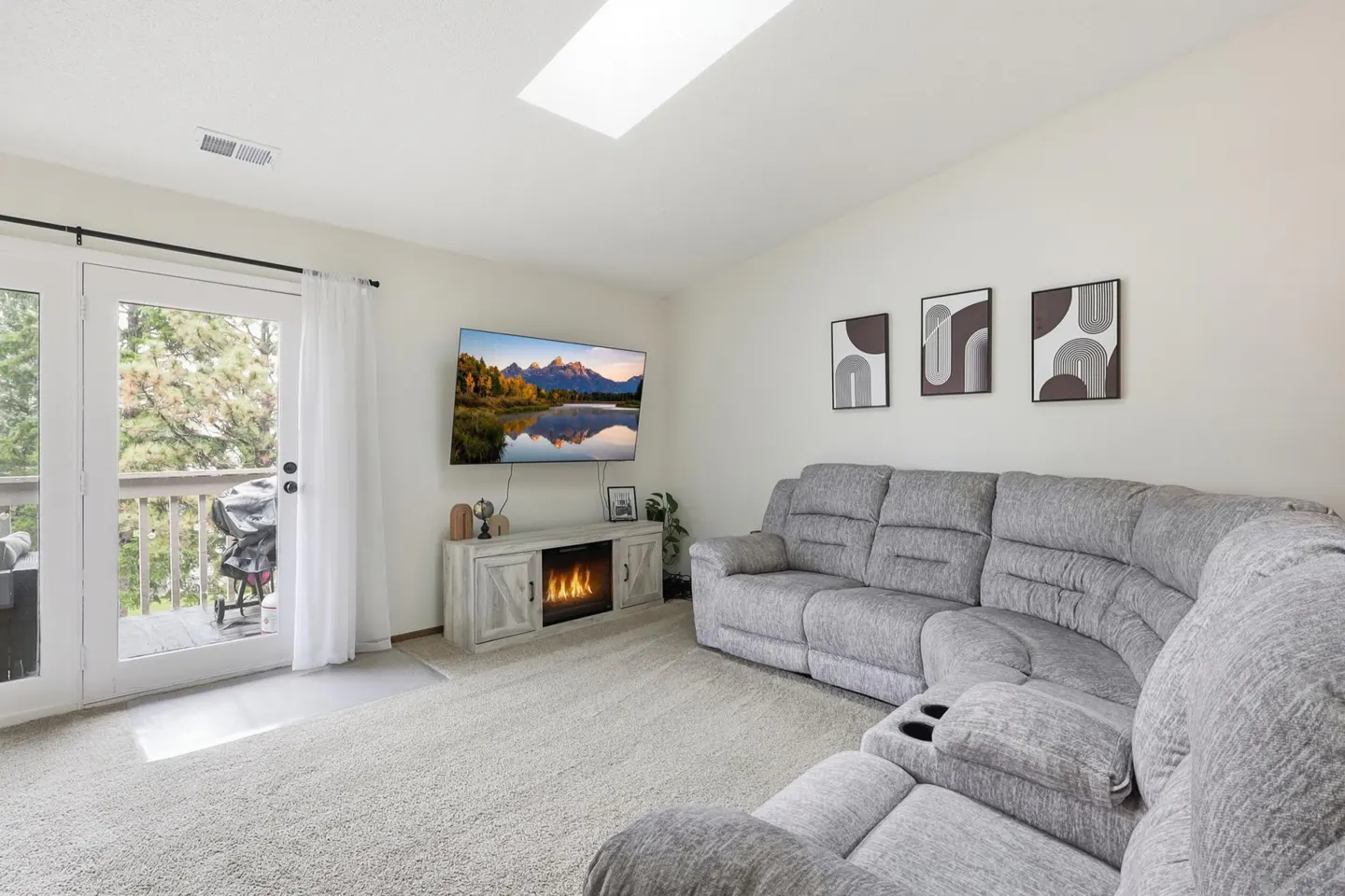 Living room with gray sectional sofa, TV above fireplace, and balcony access. Neutral tones, modern art, and a bright, airy feel.