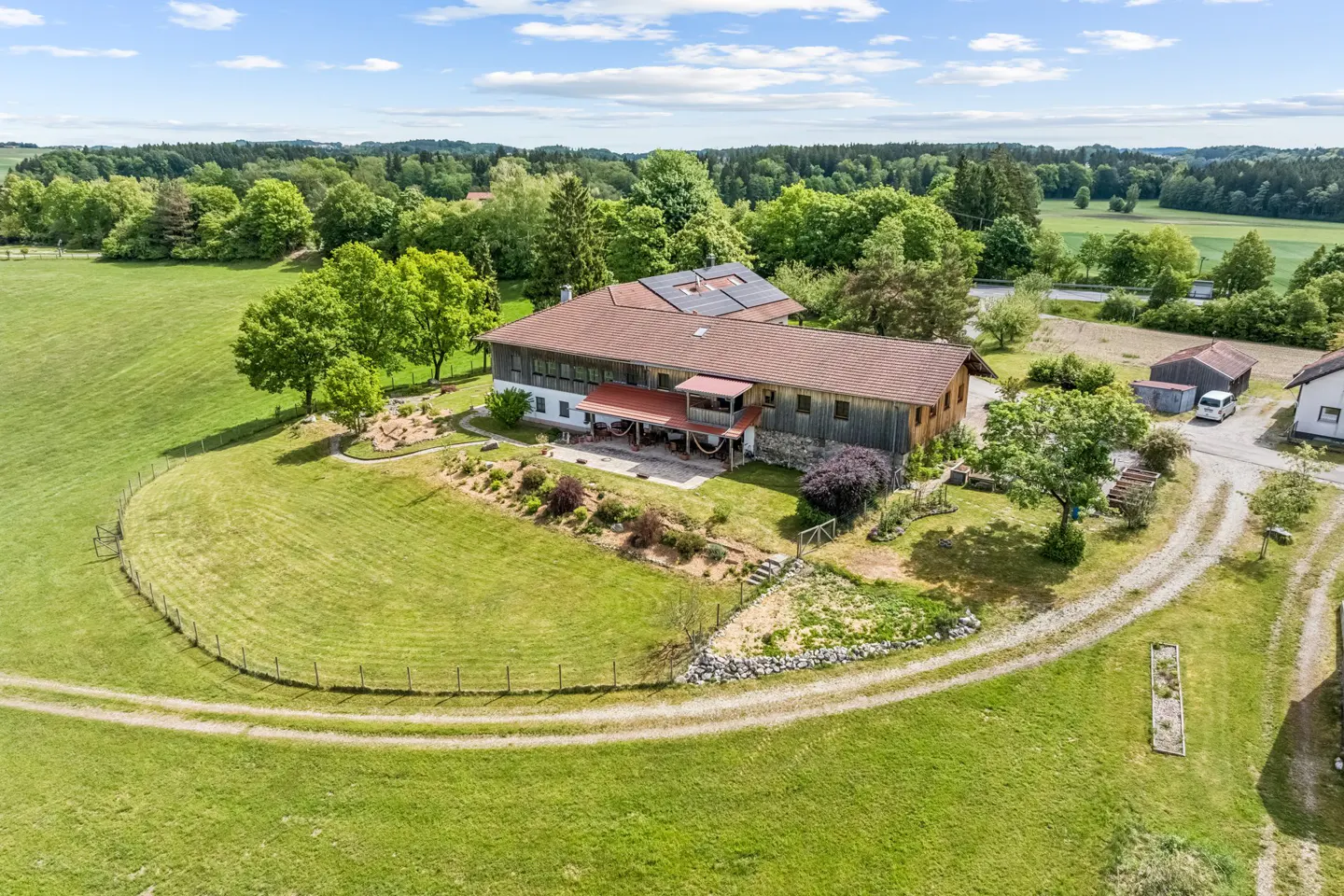 Aerial view of a rustic farmhouse with a brown roof, solar panels, and a large garden, set in a lush green rural landscape.