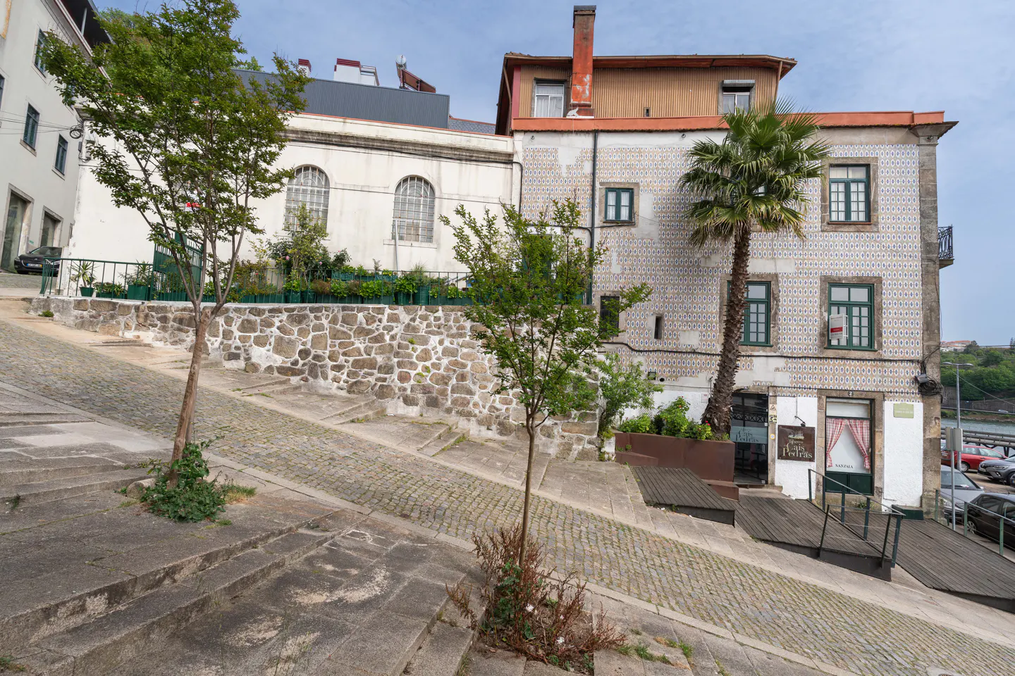 Exterior view of a building with tiled facade and green window frames, next to a cobblestone street with stairs and trees.