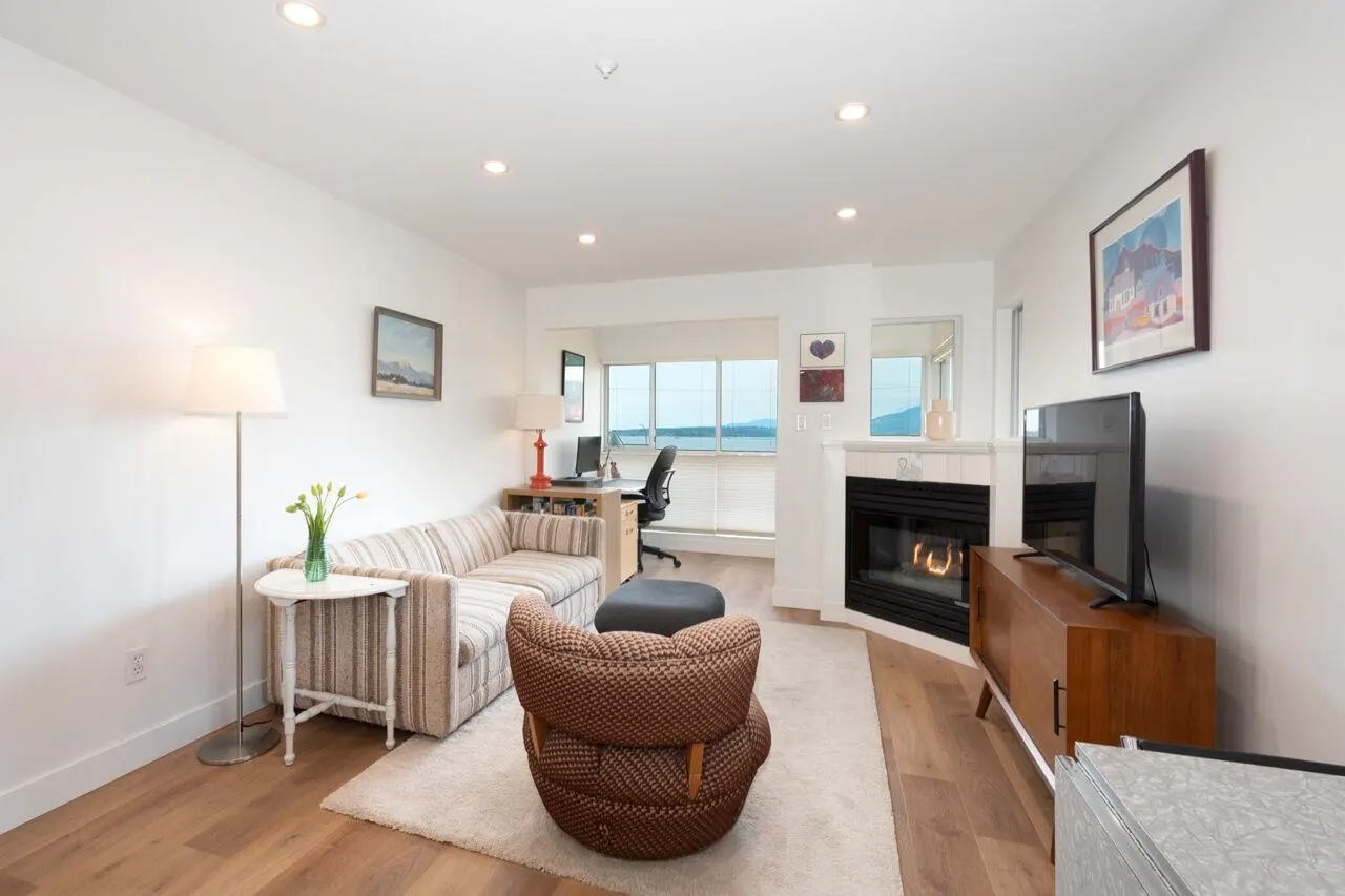 Bright living room with white walls, wood floors, and a fireplace. A striped sofa, patterned chair, and TV create a cozy space.