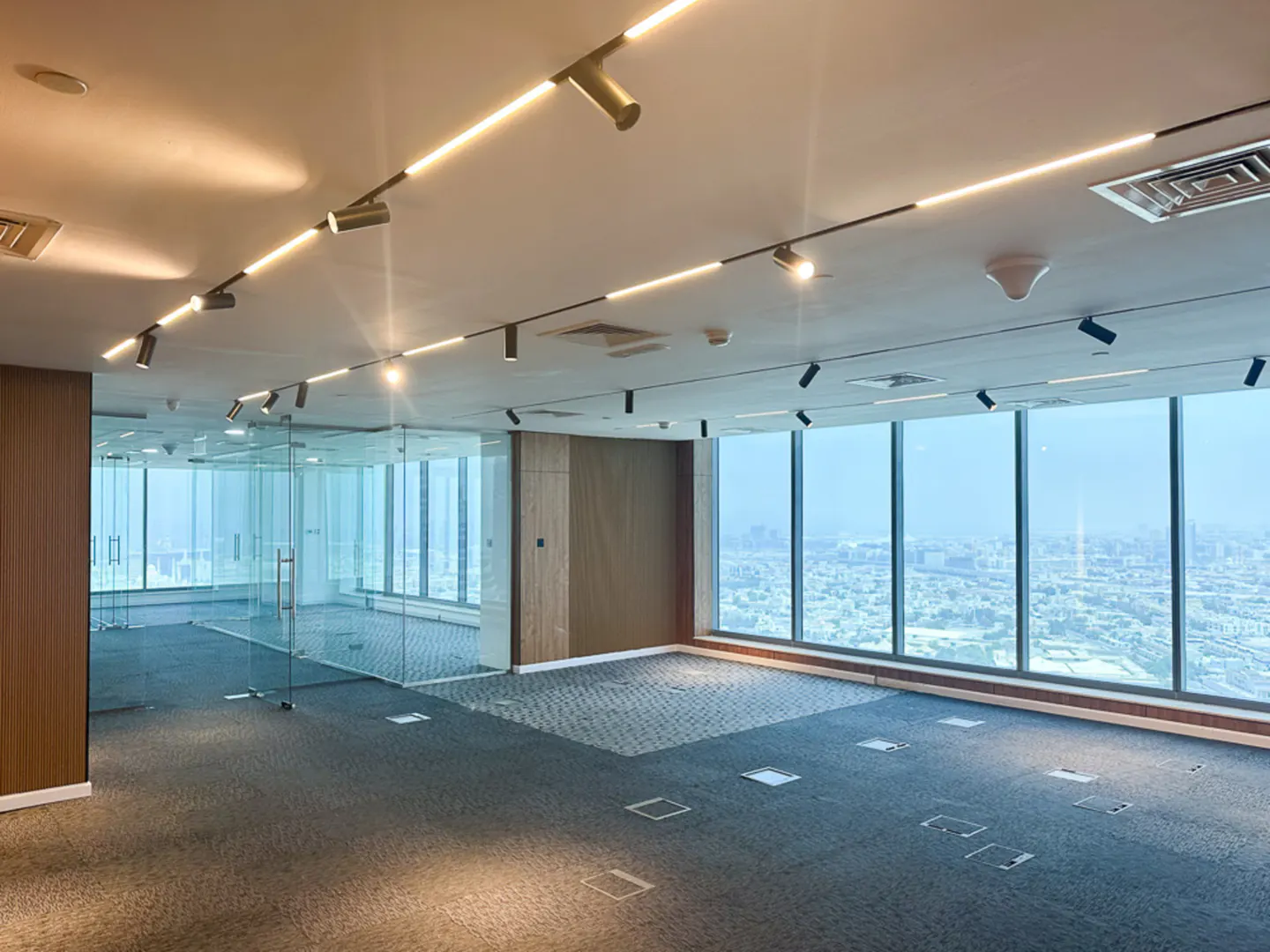 Bright, empty office space with large windows overlooking a city. Track lighting illuminates the gray carpet and glass-walled room.