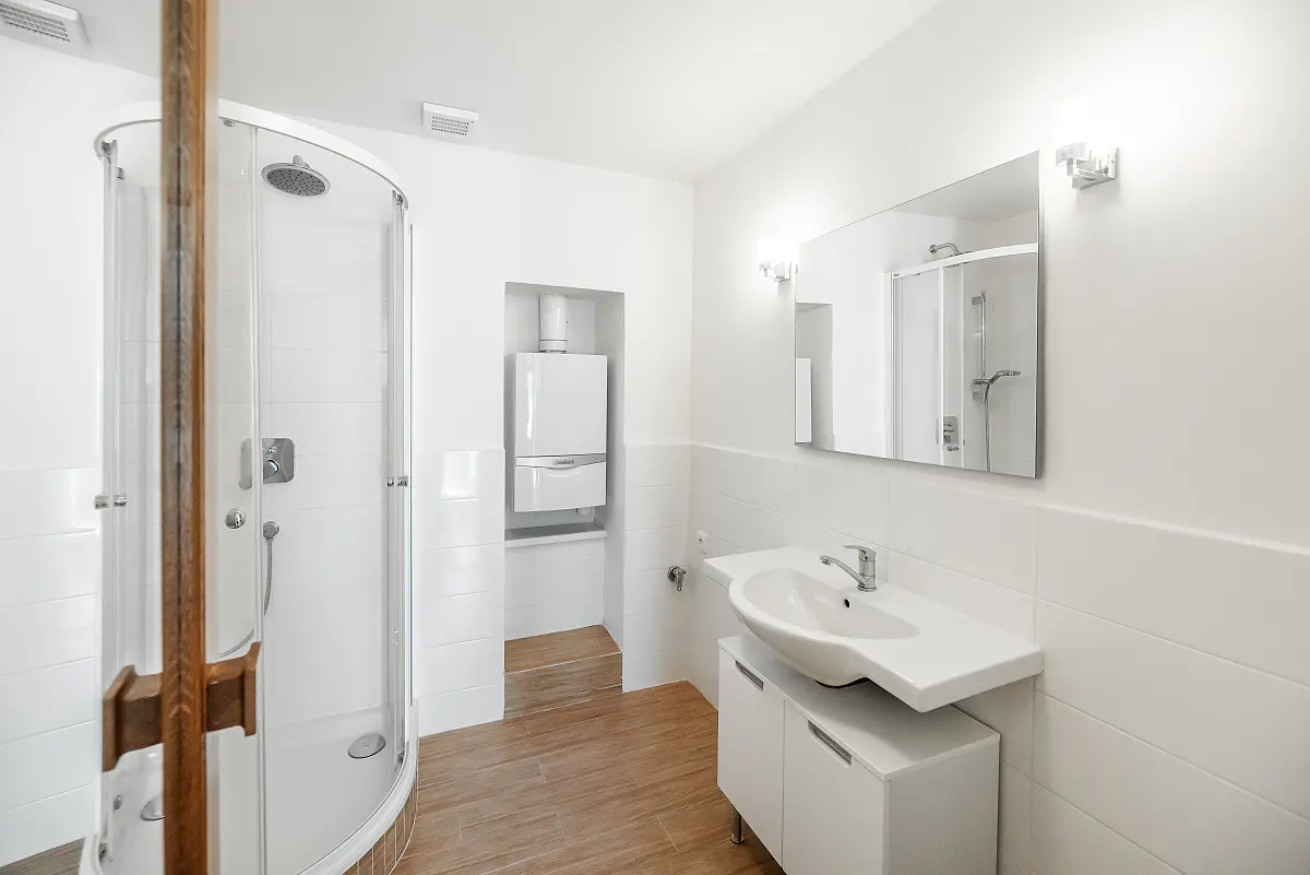 Bright bathroom with wood-look tile, white walls, and a glass shower. A white sink and cabinet are next to a recessed water heater.