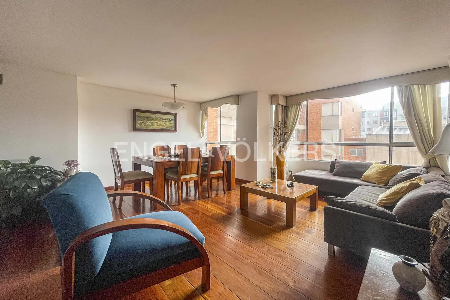 Bright living room with wood floors, dining table, gray sofa, and blue chair. Large windows offer city views.