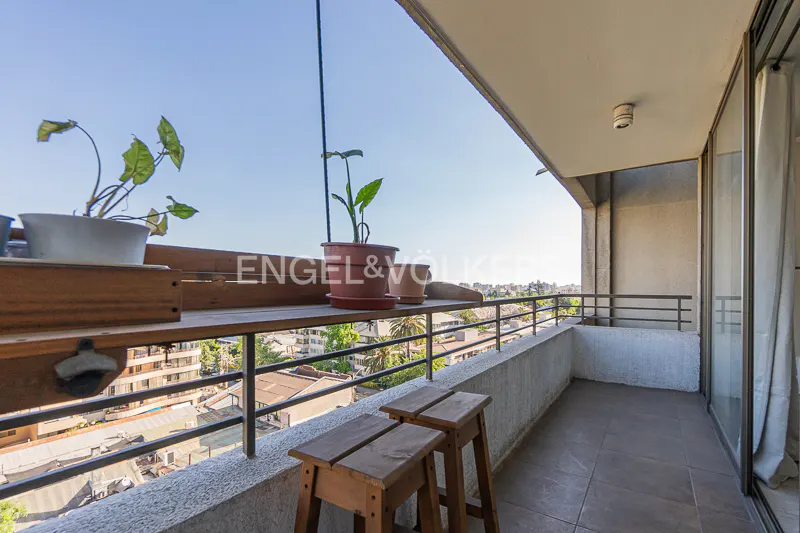 Balcony view with plants, wooden stools, and Engel & Völkers logo. Cityscape visible in the background.