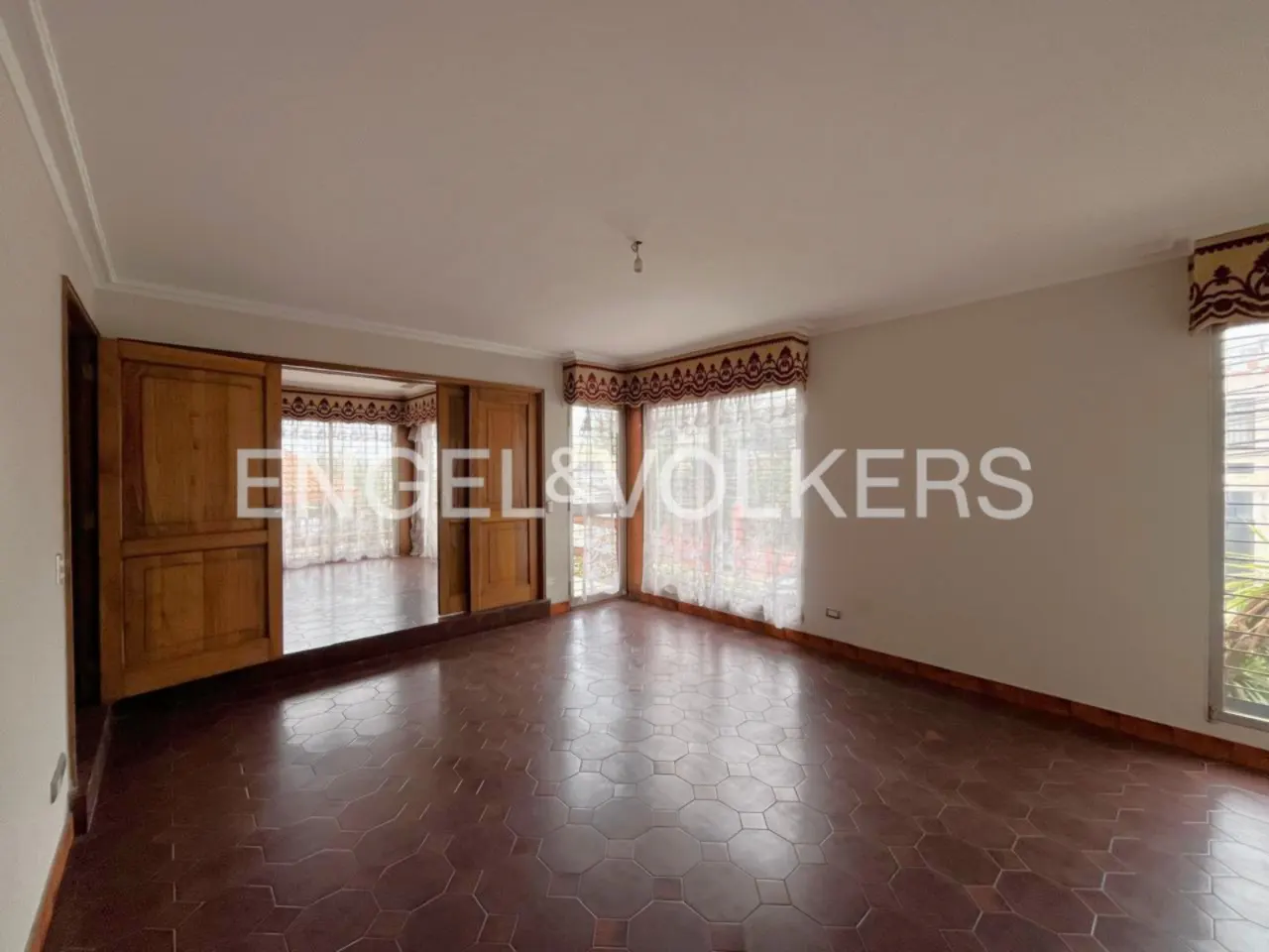 Empty living room with brown tile floor, white walls, and wooden sliding doors to another room. Windows with sheer curtains let in natural light.