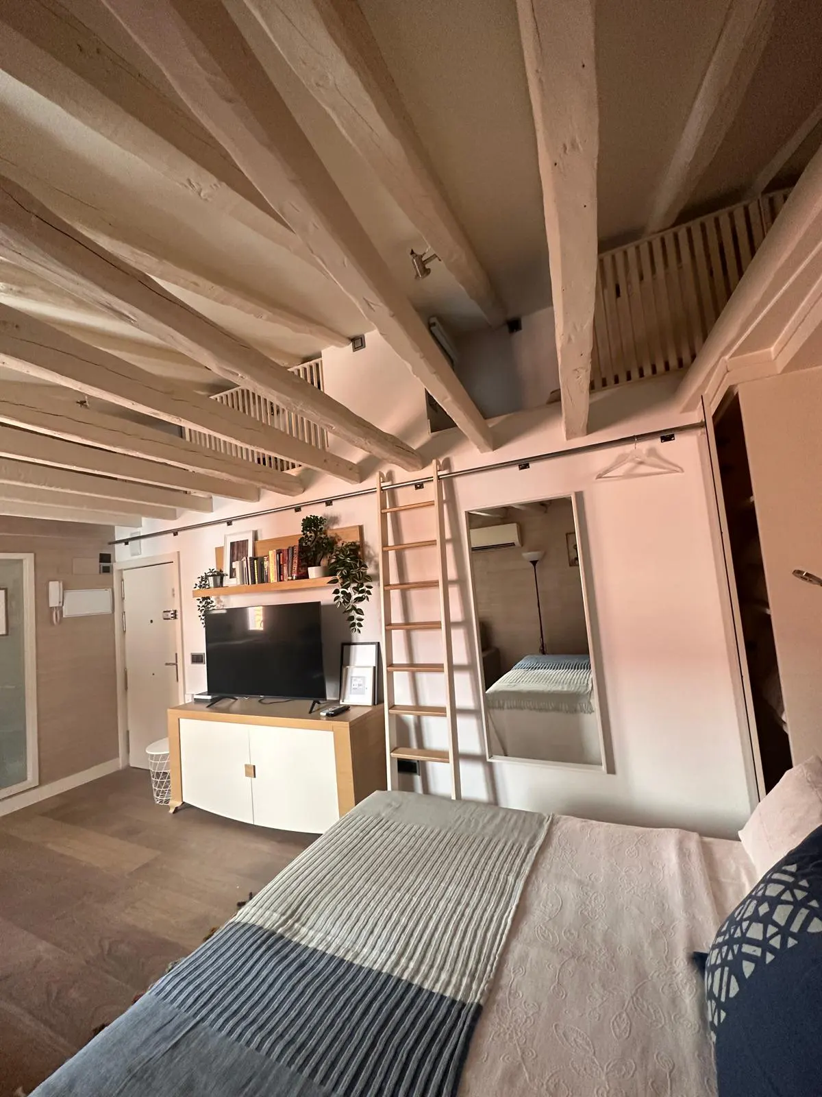 Bedroom with white exposed ceiling beams, a bed, a TV console with shelves, a ladder, and a large wall mirror.