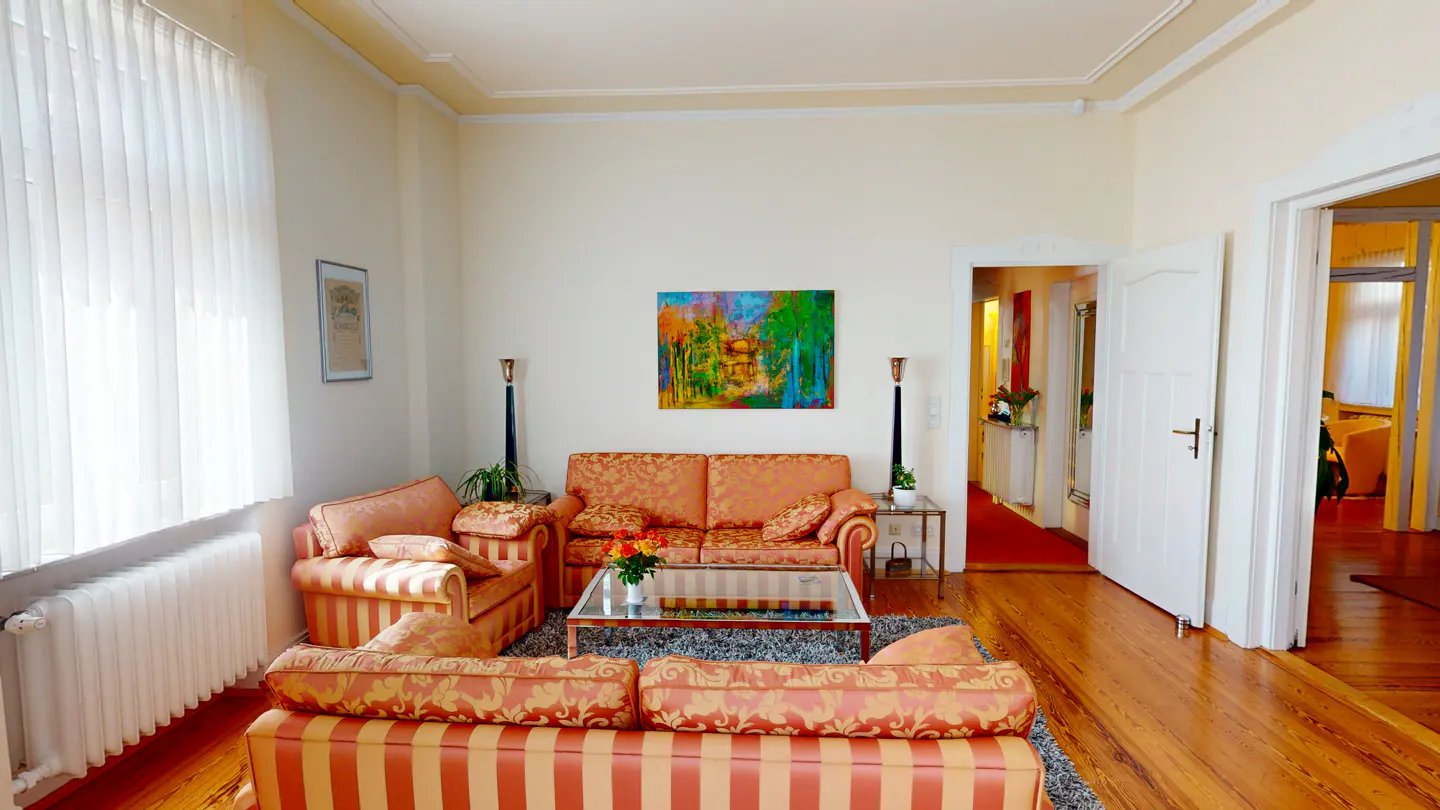 Living room with orange striped sofas, a glass table, and a colorful painting on the wall. Hardwood floors and white walls.