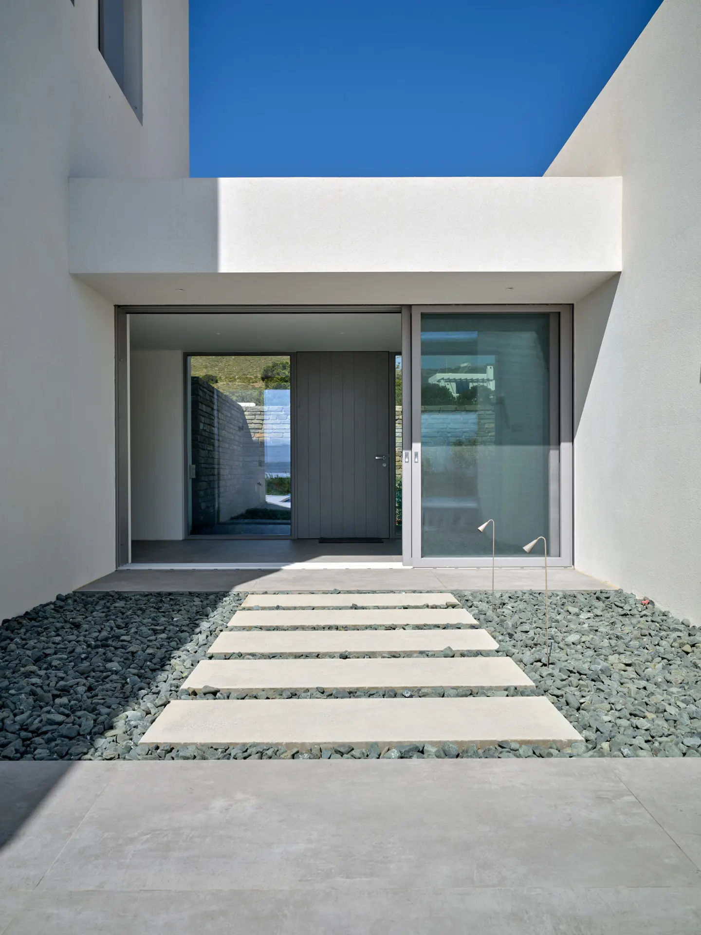 Modern home entrance with a gray door, glass window, and stone pathway leading to the entrance. The walls are white, and the sky is blue.