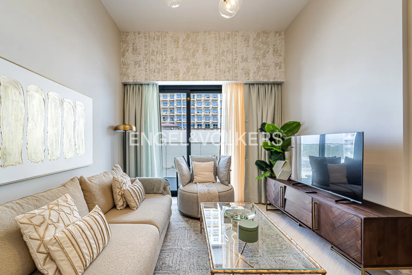 Living room with beige sofa, round chair, glass coffee table, and TV stand. Balcony doors with curtains in the background.