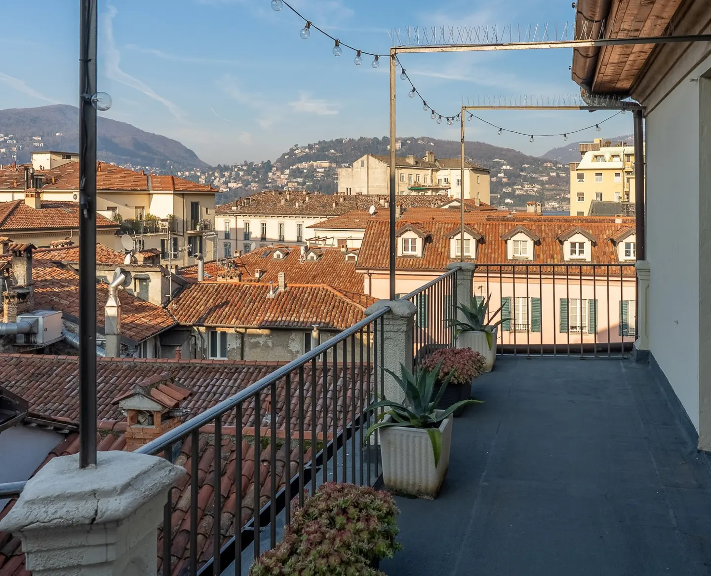View from a rooftop terrace with potted plants, black metal railing, and red tile rooftops in the background.