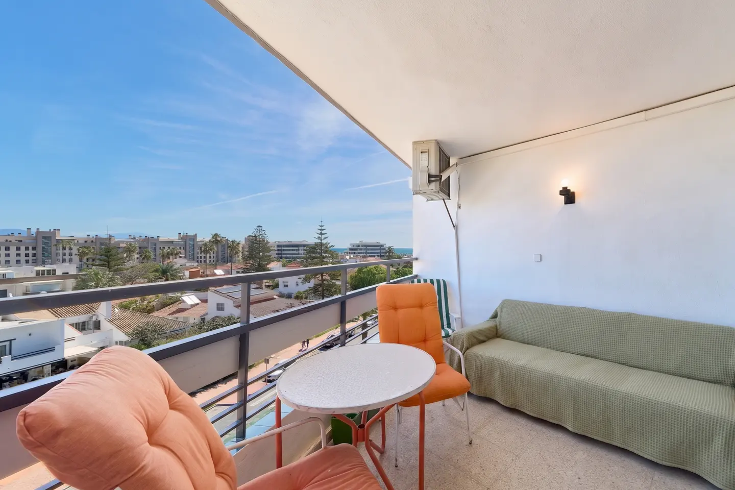 Balcony view with seating. Orange chairs surround a white table. A green sofa sits against a white wall. City and ocean views in the background.