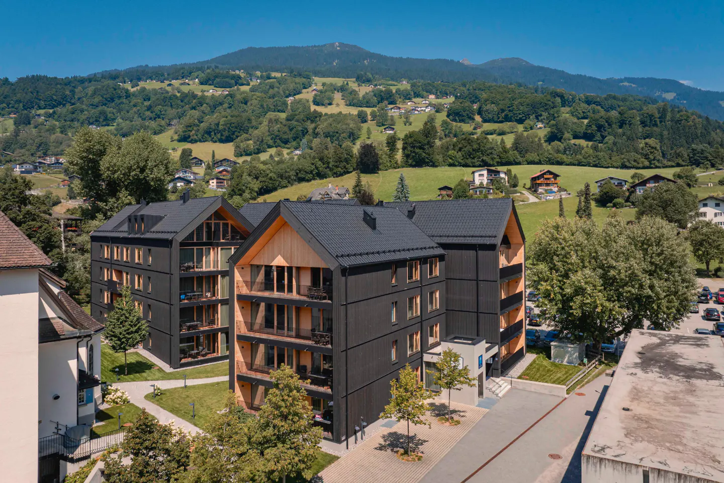 Modern black apartment buildings with wooden balconies set against a lush green mountain landscape under a clear blue sky.