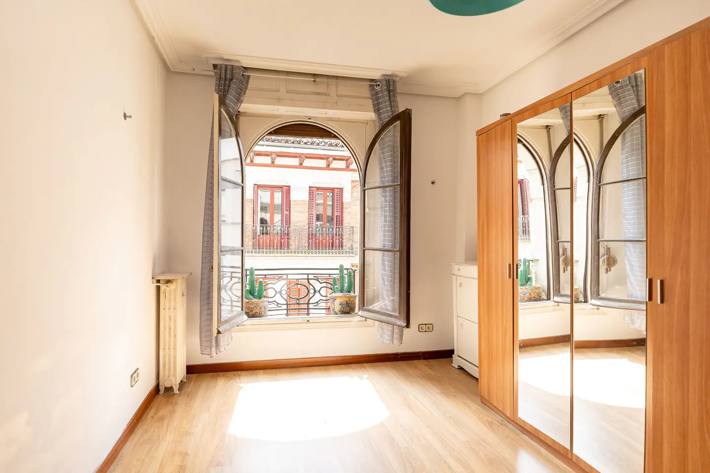A bright bedroom with a large arched window, wood floors, and a mirrored wardrobe.