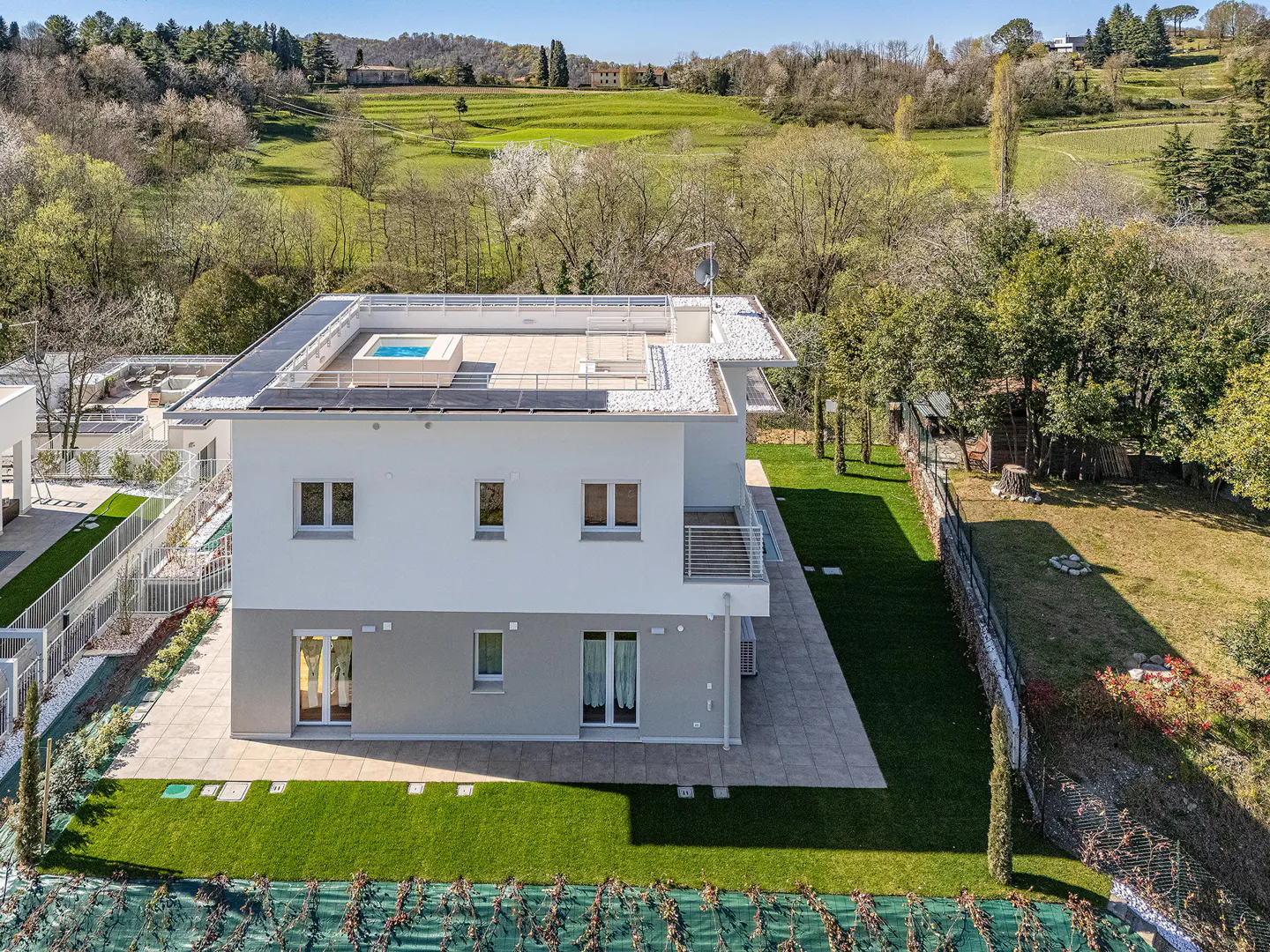 Aerial view of a modern two-story house with a rooftop pool and solar panels, surrounded by green lawns and trees.