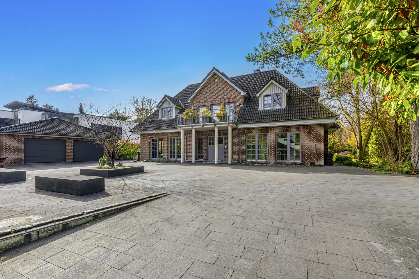 Brick house with a gray roof, balcony with yellow flowers, and a large paved driveway under a blue sky.