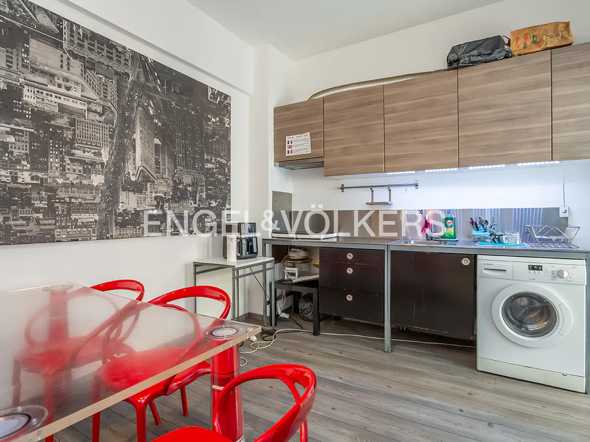 A kitchen with a red table and chairs, gray cabinets, and a black and white city print on the wall. A washing machine is visible.