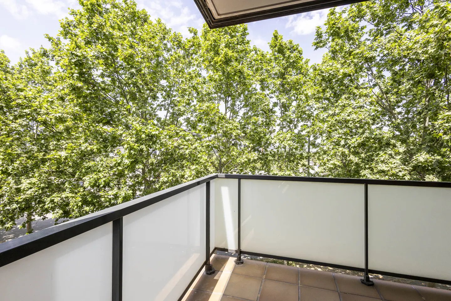 Balcony view with black railing and frosted glass panels. Green trees fill the background under a blue sky. Brown tiled floor.