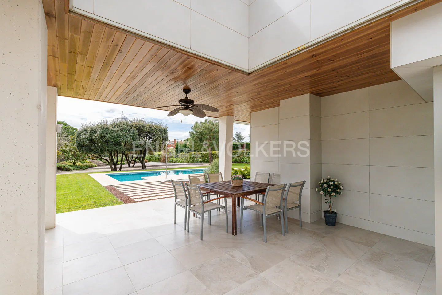 Covered patio with wood ceiling, fan, and stone floor. Table and chairs overlook a pool and green lawn.