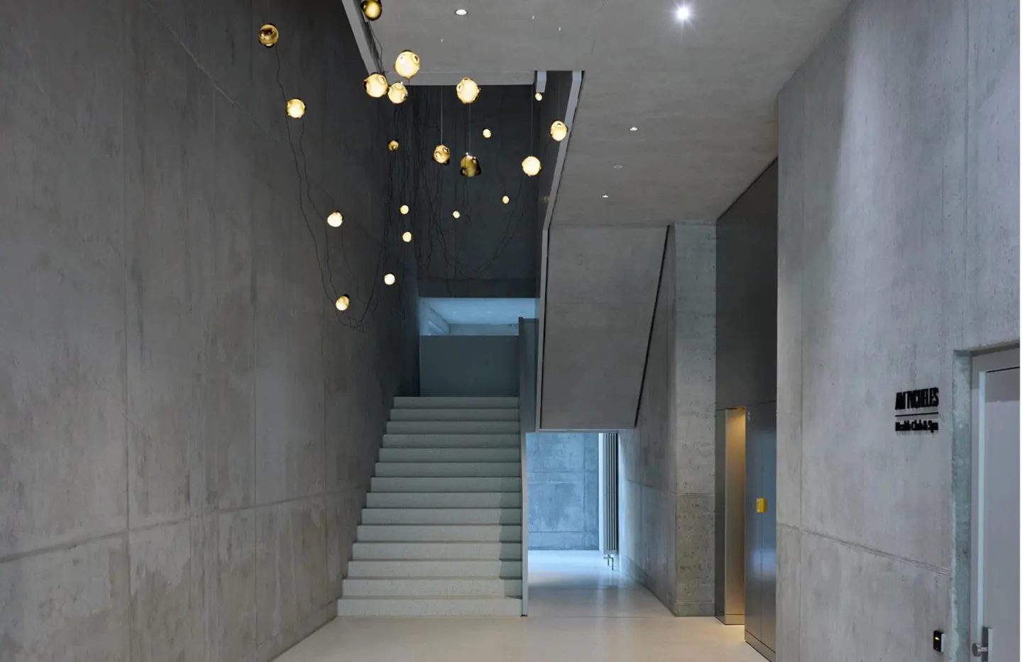 Modern gray concrete lobby with white stairs, elevator, and globe pendant lights.