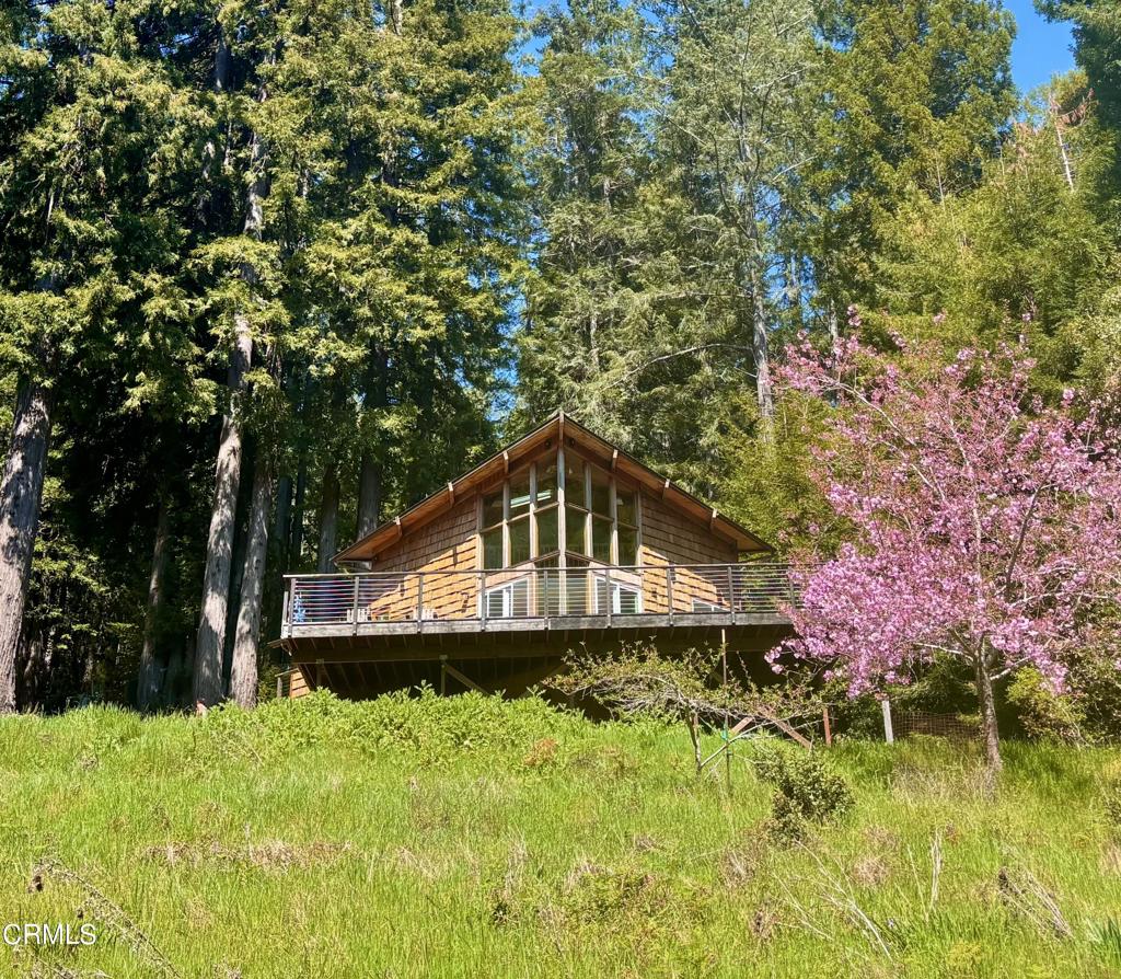 A wood-shingled A-frame house with a deck sits on a grassy hill surrounded by tall trees and a pink flowering tree.