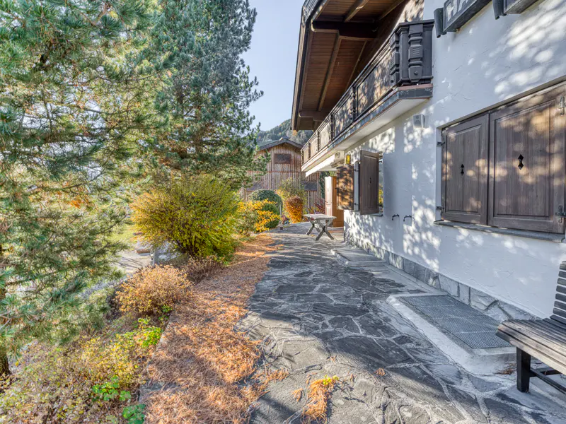 Exterior view of a white house with brown shutters and a balcony, surrounded by trees and a stone patio.