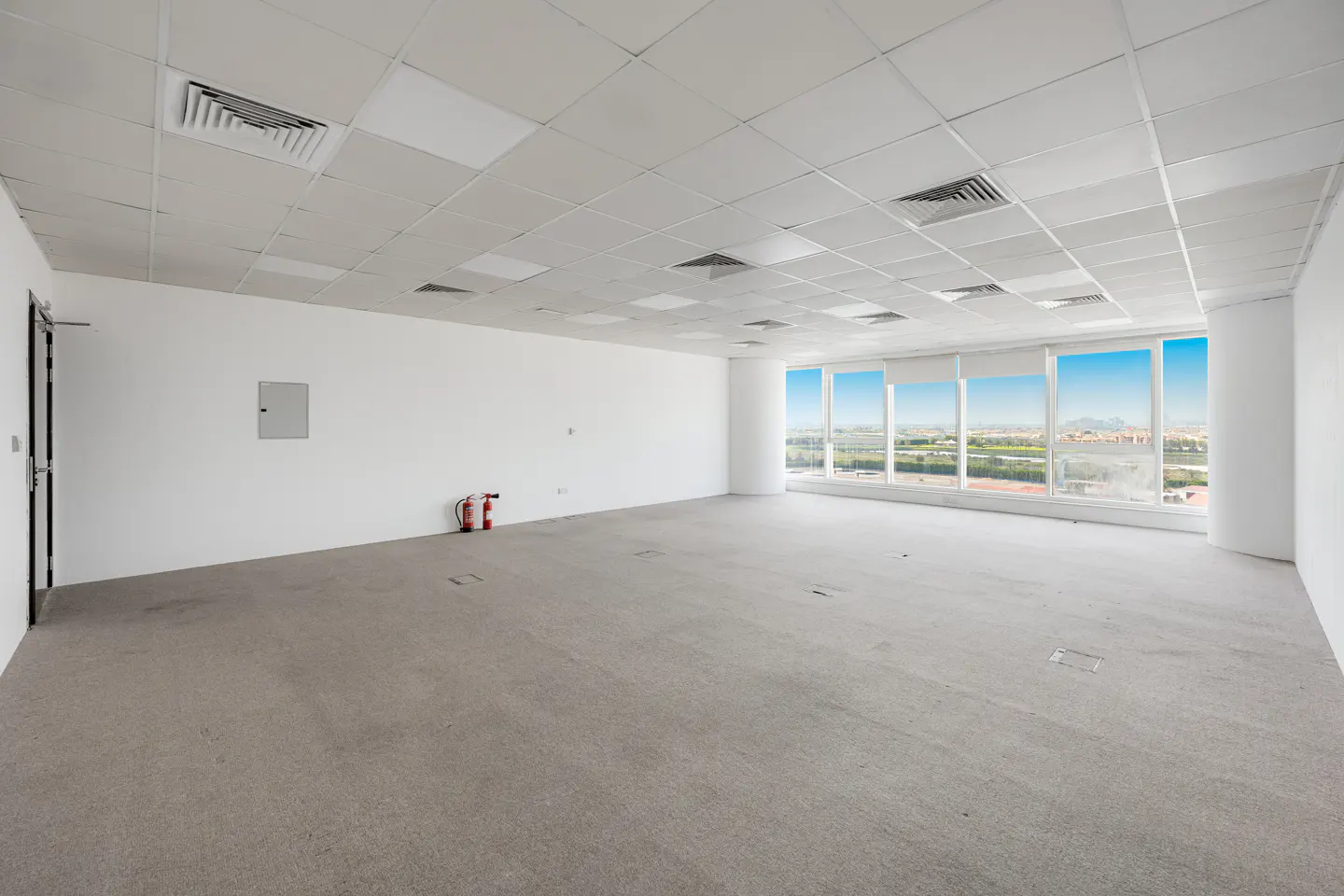 Empty office space with white walls, gray carpet, and large windows showing a city view. A fire extinguisher stands near the wall.