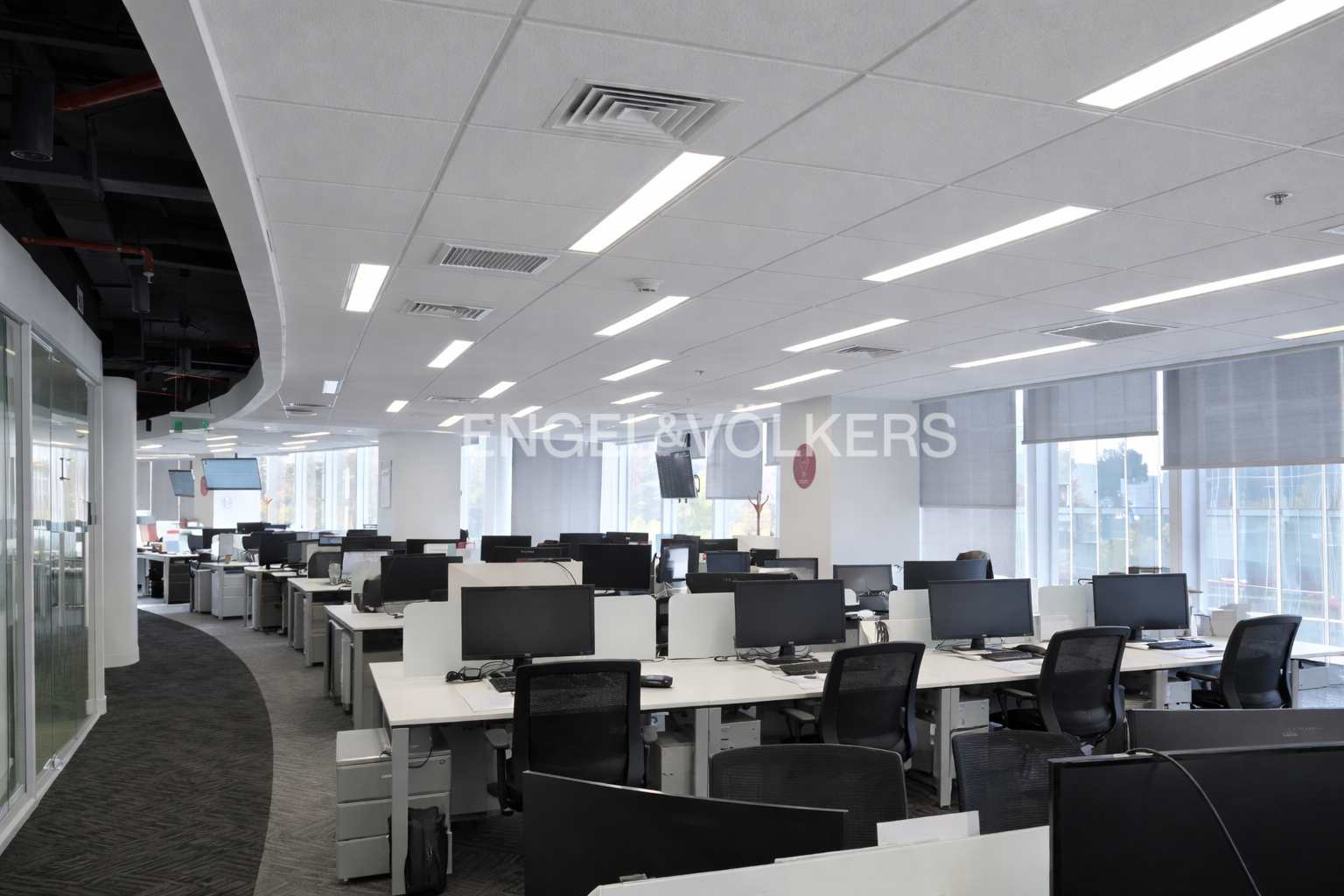 Bright, modern office space with rows of white desks and black chairs. Large windows provide natural light. Engel & Volkers logo visible.