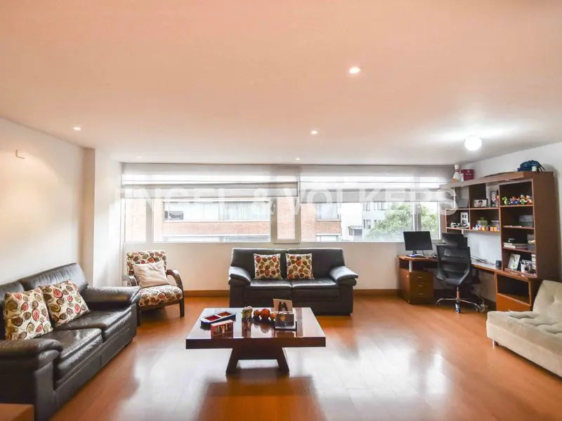Bright living room with hardwood floors, two dark leather sofas, patterned armchair, and a dark wood coffee table. A desk and shelves are visible in the background.