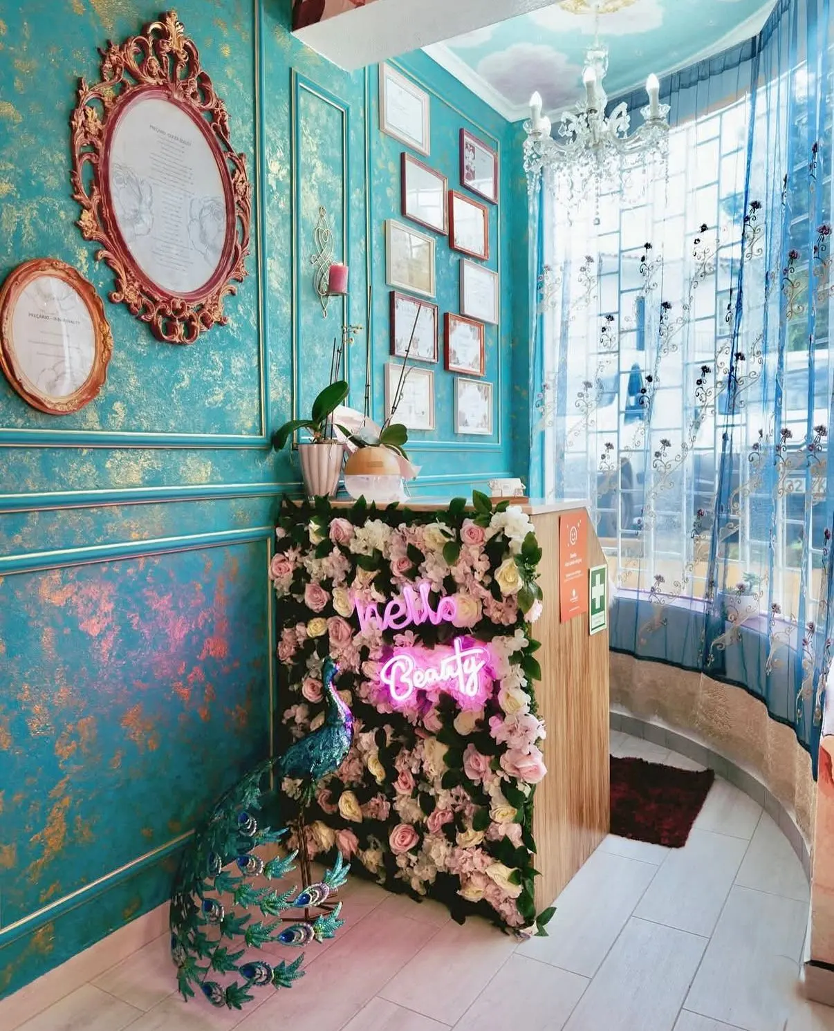 Interior view of a beauty salon with turquoise walls, ornate frames, a flower-covered counter with a neon "Hello Beauty" sign, and a peacock statue.