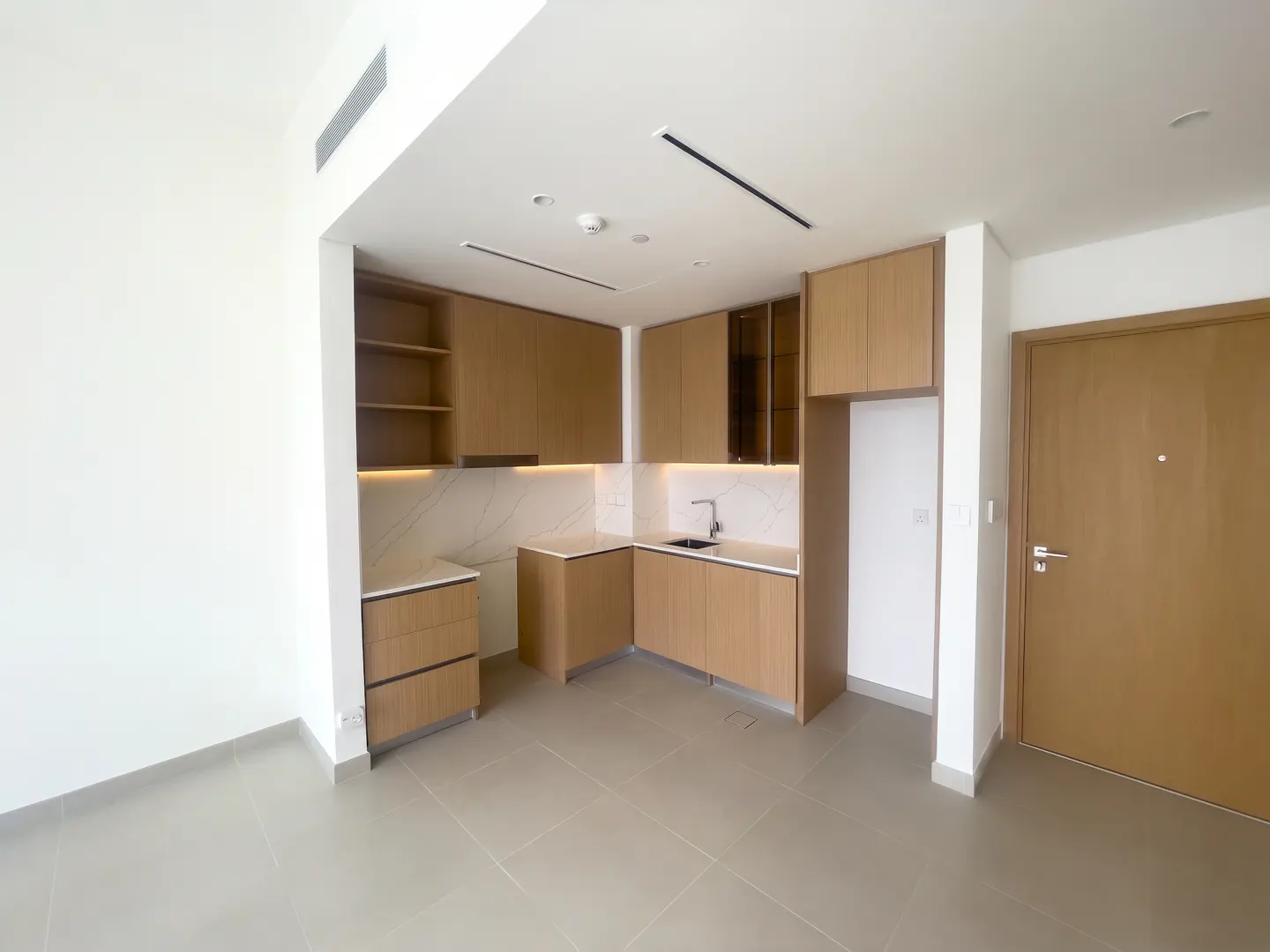 A bright, modern kitchen with light wood cabinets, white marble backsplash, and gray tile flooring. A closed door is visible on the right.