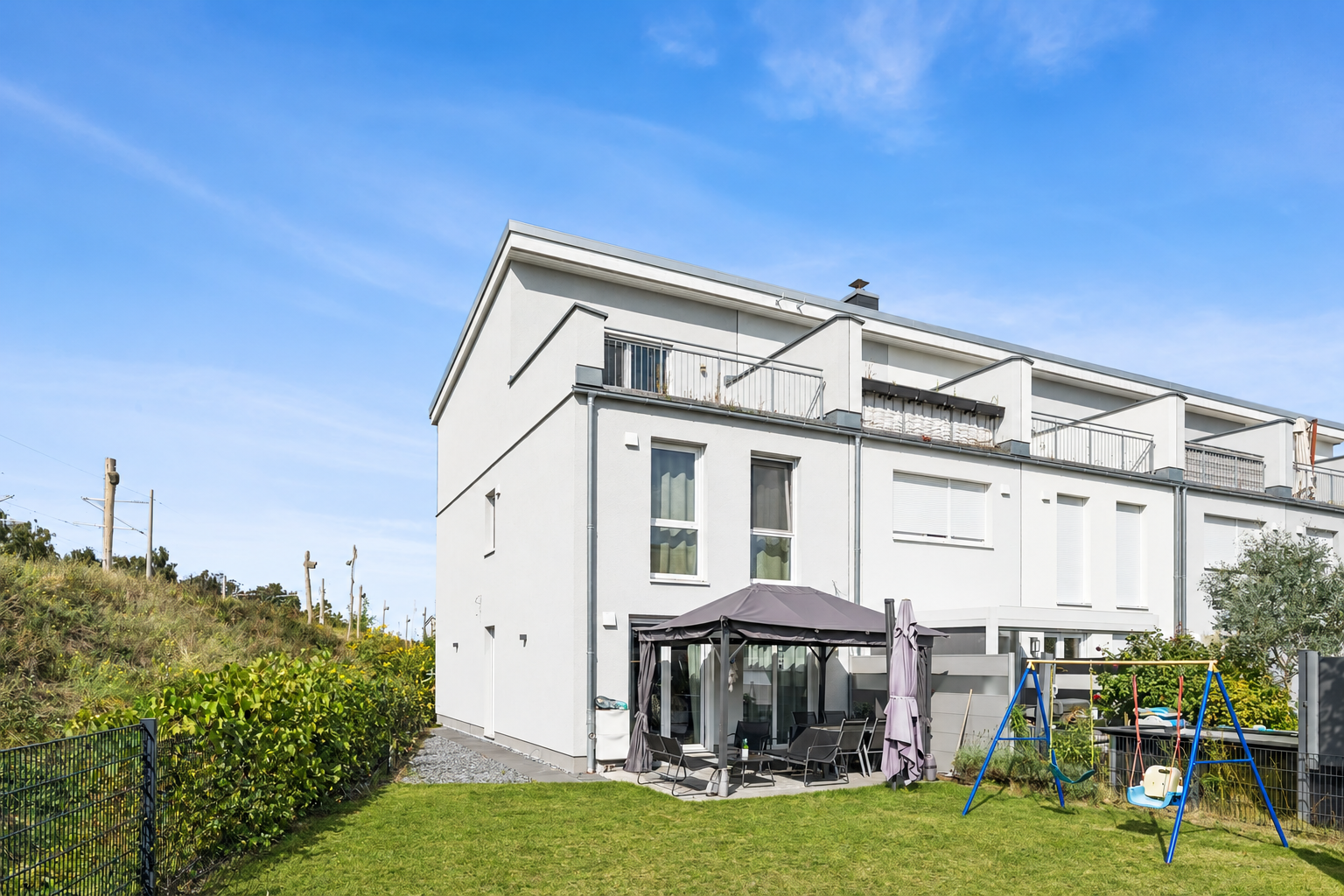 Backyard of a modern white townhouse with a patio, gazebo, and a blue swing set on a green lawn under a clear blue sky. [AI-generated text]