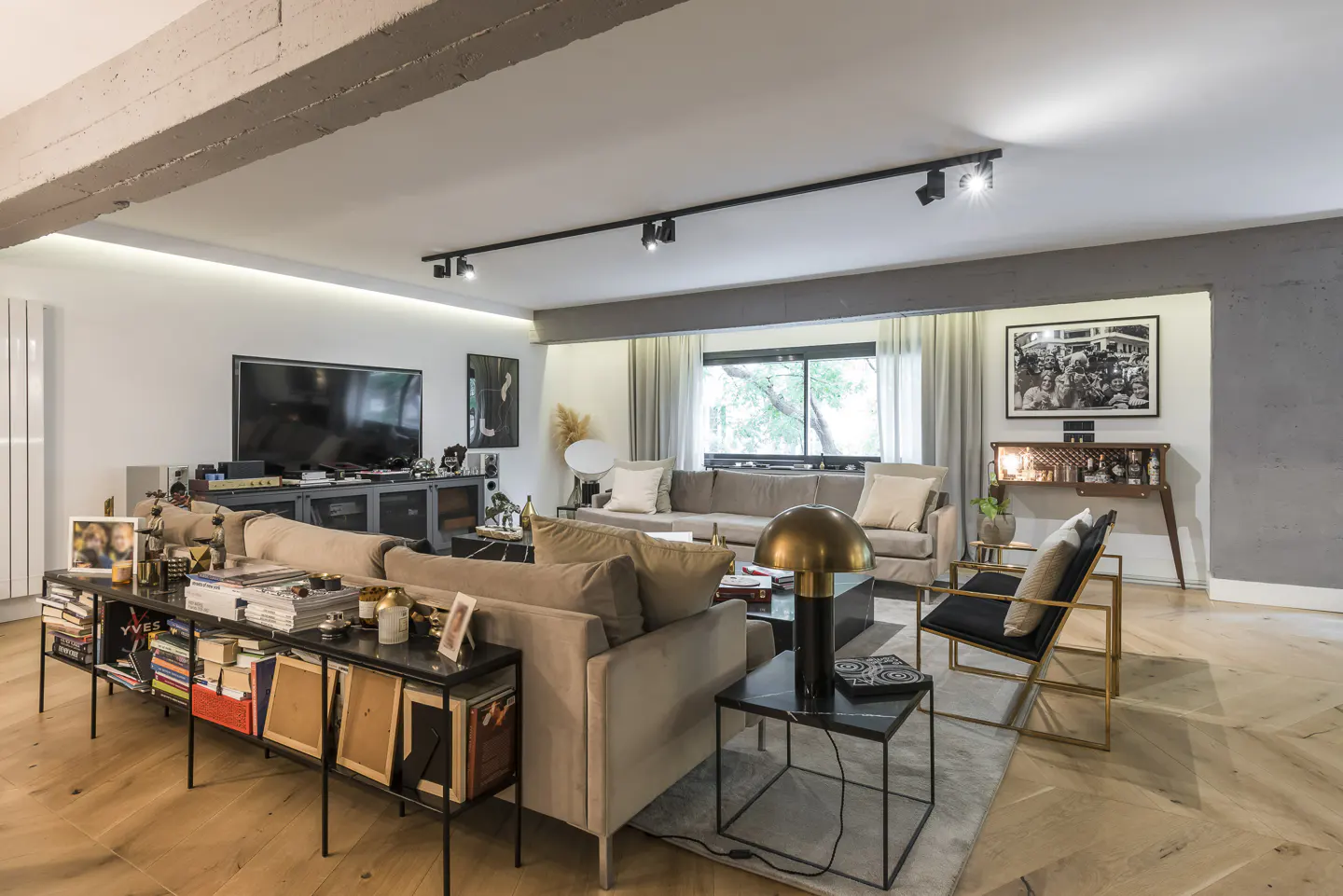 A modern living room with beige sofas, black tables, and a console table filled with books and frames. A TV and bar area are in the background.