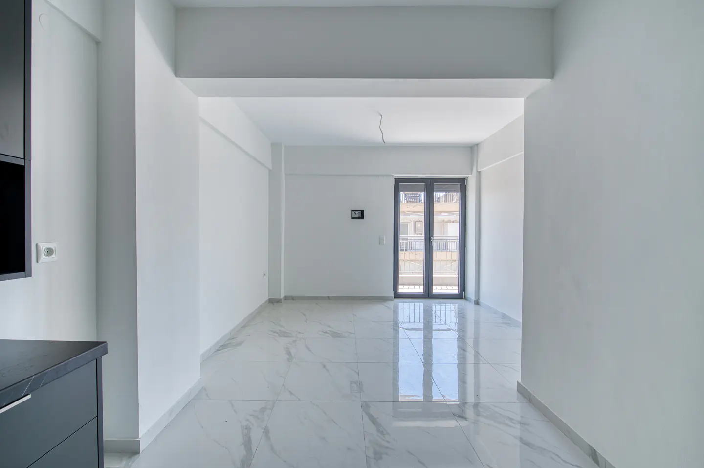 Bright, empty room with white walls and marble floors. A gray door leads to a balcony with a city view. A dark cabinet is on the left.