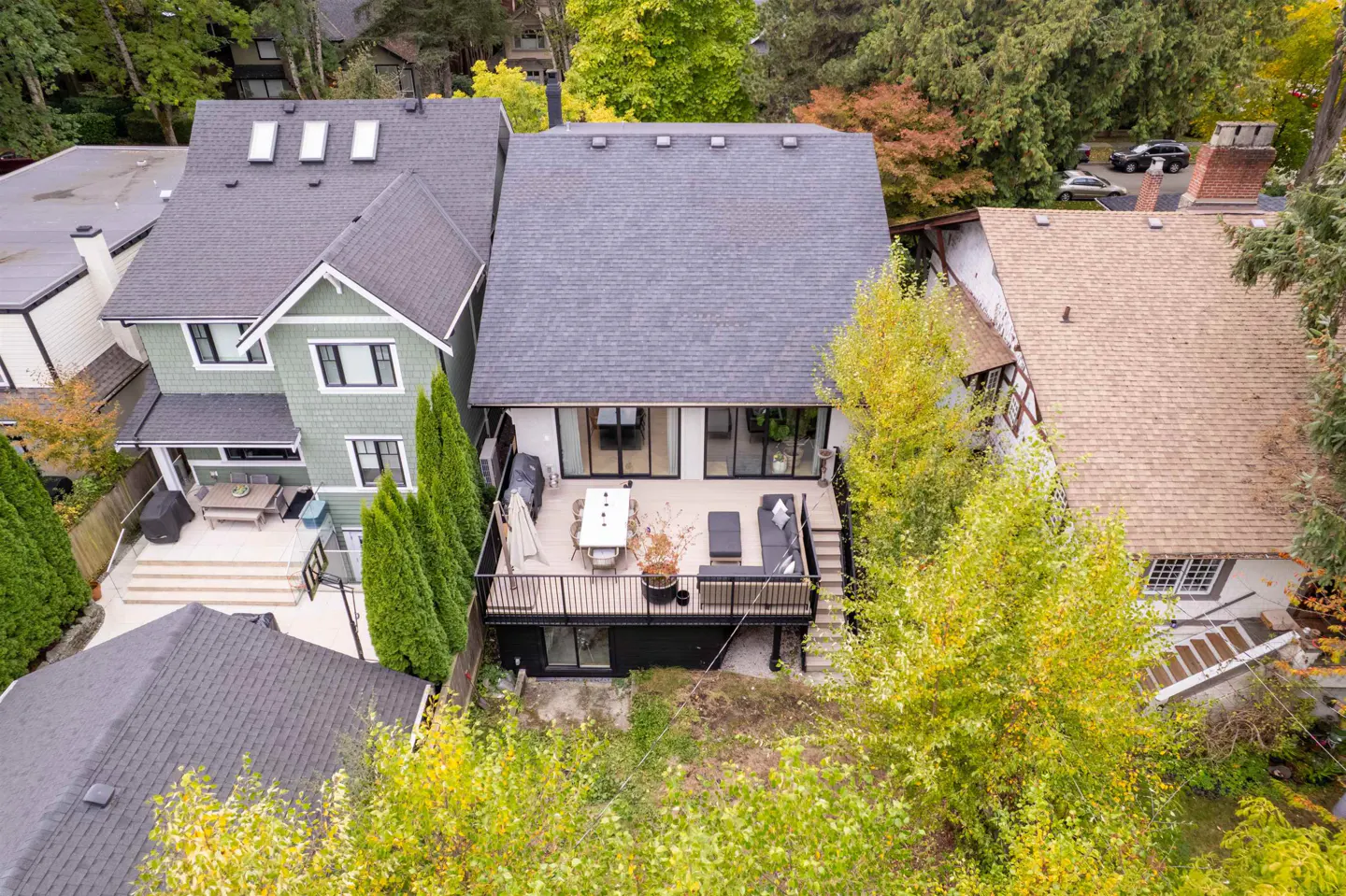 Aerial view of a modern house with a gray roof and a large deck with outdoor furniture. Green trees surround the house.
