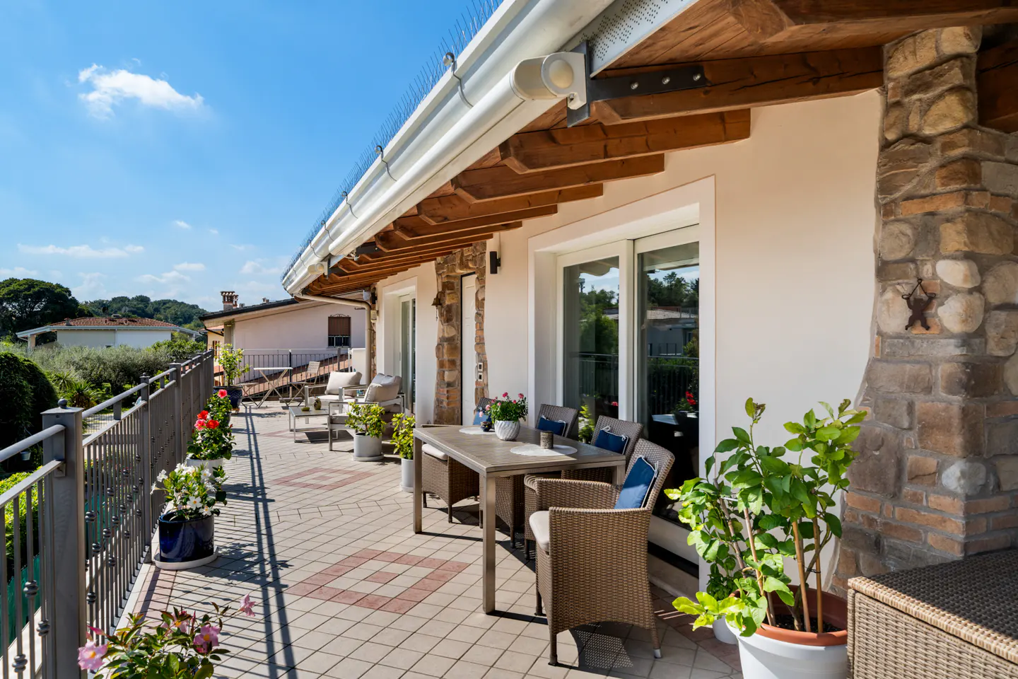 A sunny balcony with a dining table, chairs, and potted plants. The balcony has a stone accent wall and overlooks a green landscape.