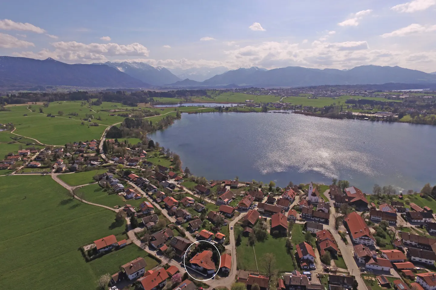 Aerial view of a lakeside village with red-roofed houses, green fields, and snow-capped mountains in the background. A white circle highlights a specific house.