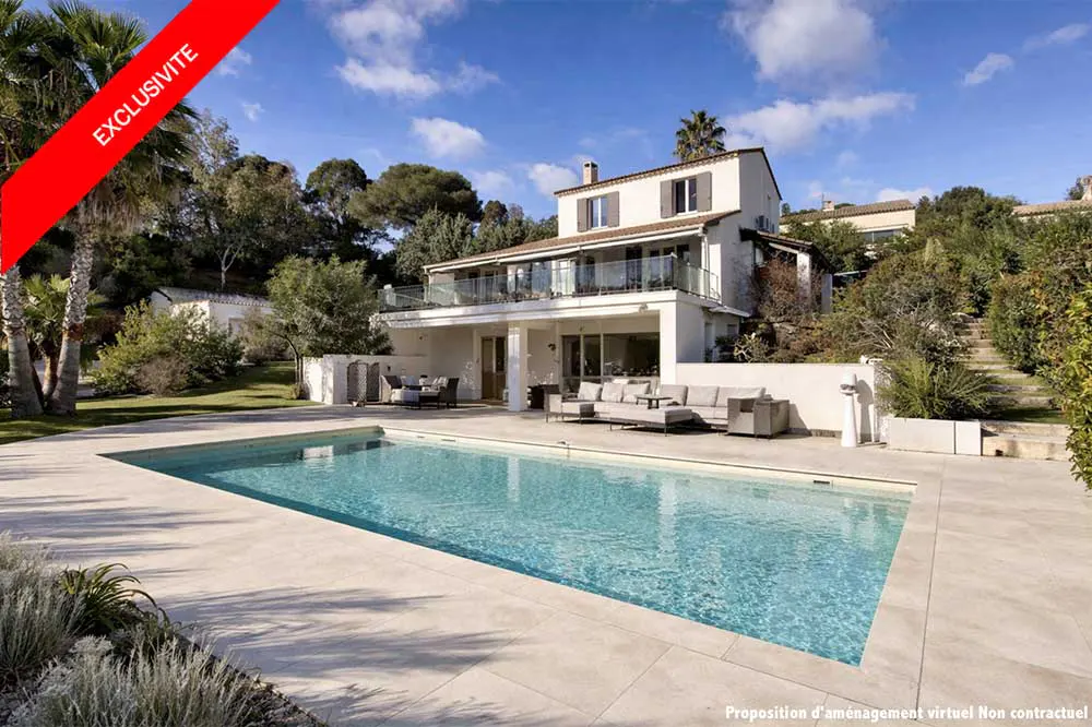 A two-story white house with a pool and patio furniture on a sunny day. A red "Exclusivite" banner is in the upper left corner.