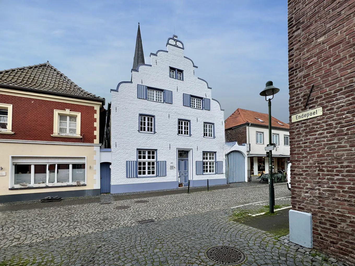 Street view of a white, gabled house with blue shutters on a cobblestone street. A brick building and street lamp are visible.