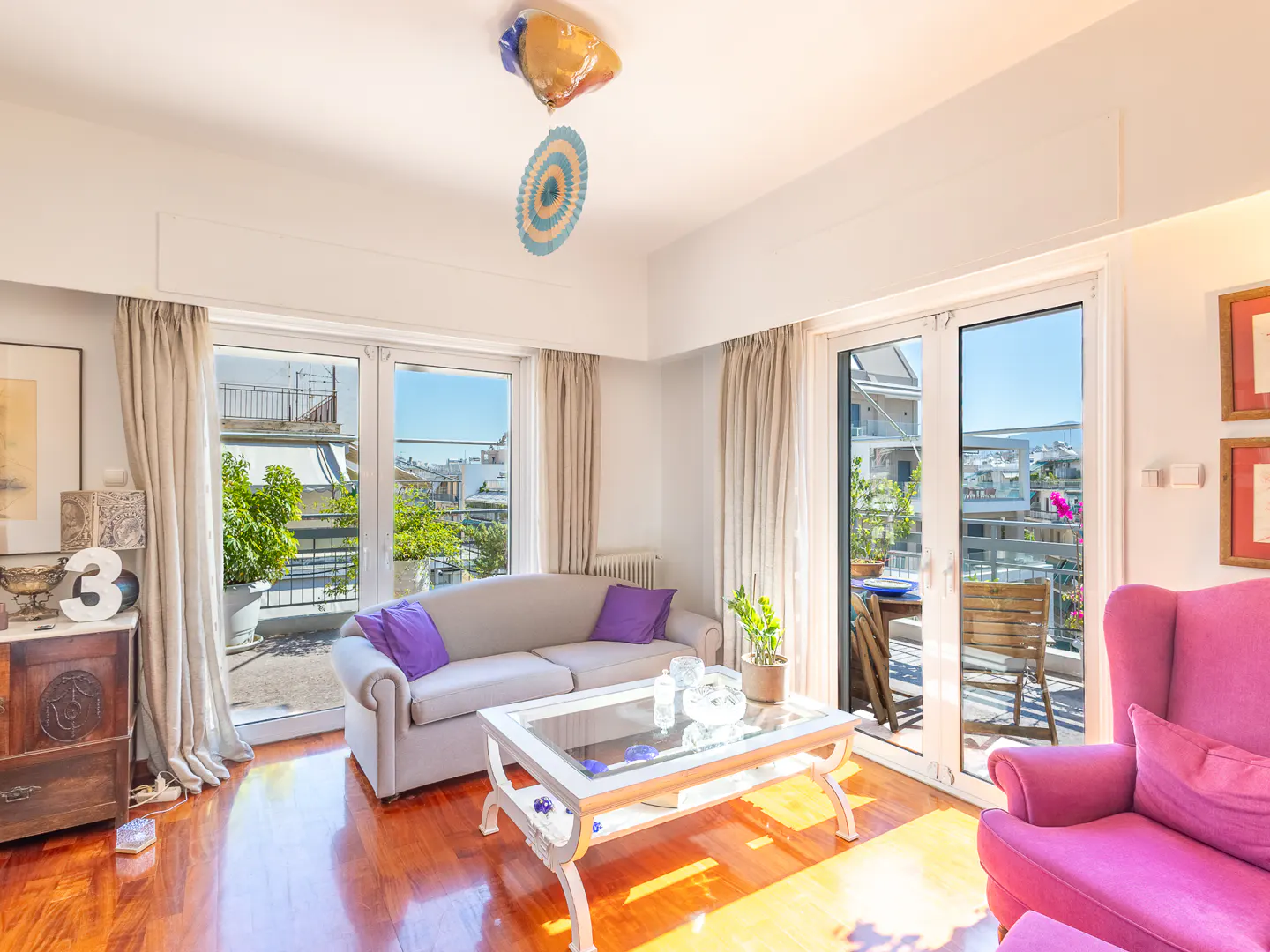 Bright living room with a light gray sofa, purple pillows, a glass coffee table, and a vibrant pink armchair.