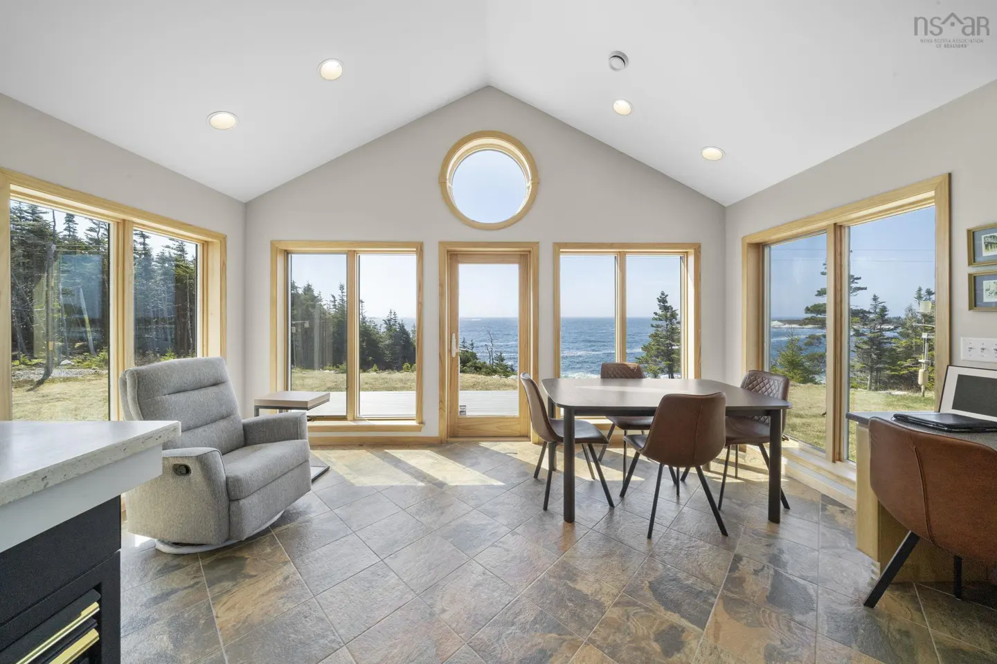 Bright sunroom with ocean view. Table, chairs, recliner, desk, and large windows. Round window above door.