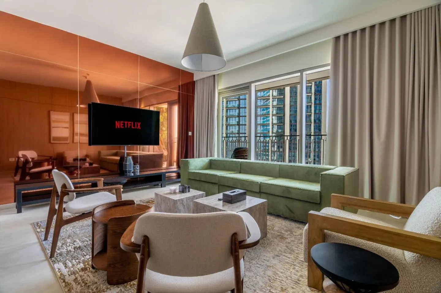 Living room with a green sofa, chairs, and a TV showing Netflix. A mirrored wall reflects the room. Balcony with city view.