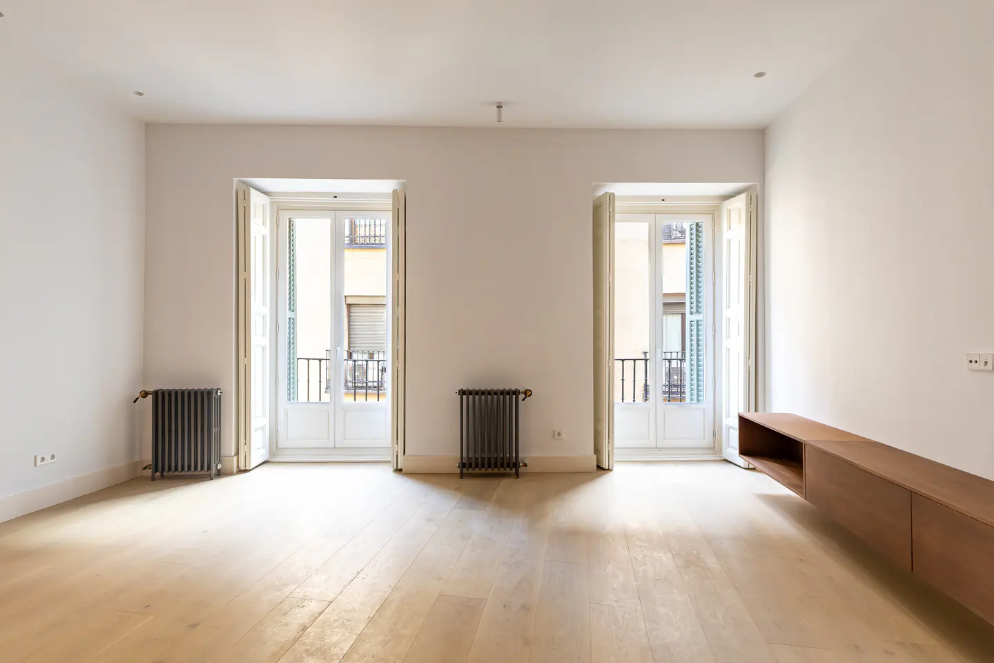 Bright, empty room with light wood floors, white walls, and two open French doors with blue shutters leading to balconies. A brown cabinet and two black radiators are visible.