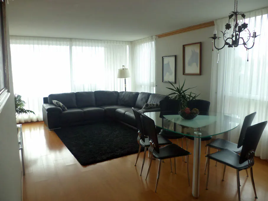 Living room with a black sectional sofa, black rug, glass table with four chairs, and sheer white curtains.